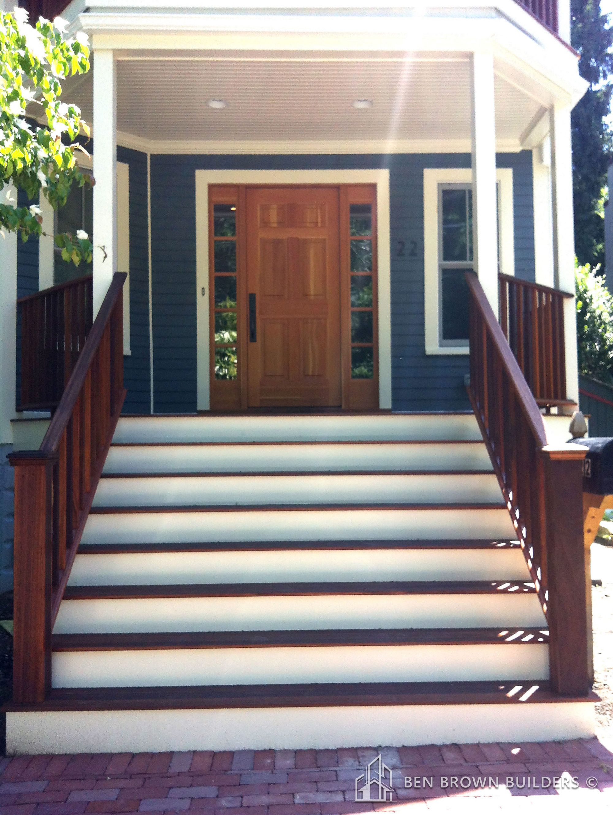 Wooden front door of a house with dark blue siding and white stairs flanked by brown railings
