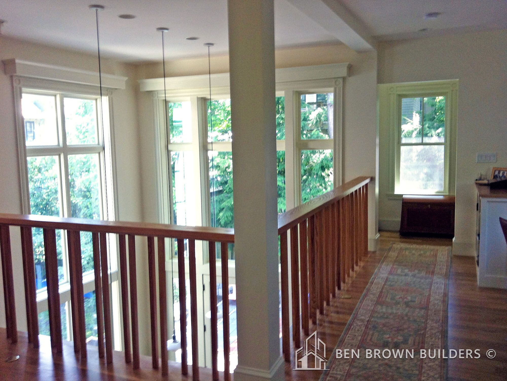 Open hallway with wooden railings, pendant lights, hardwood floors, and multiple windows with greenery