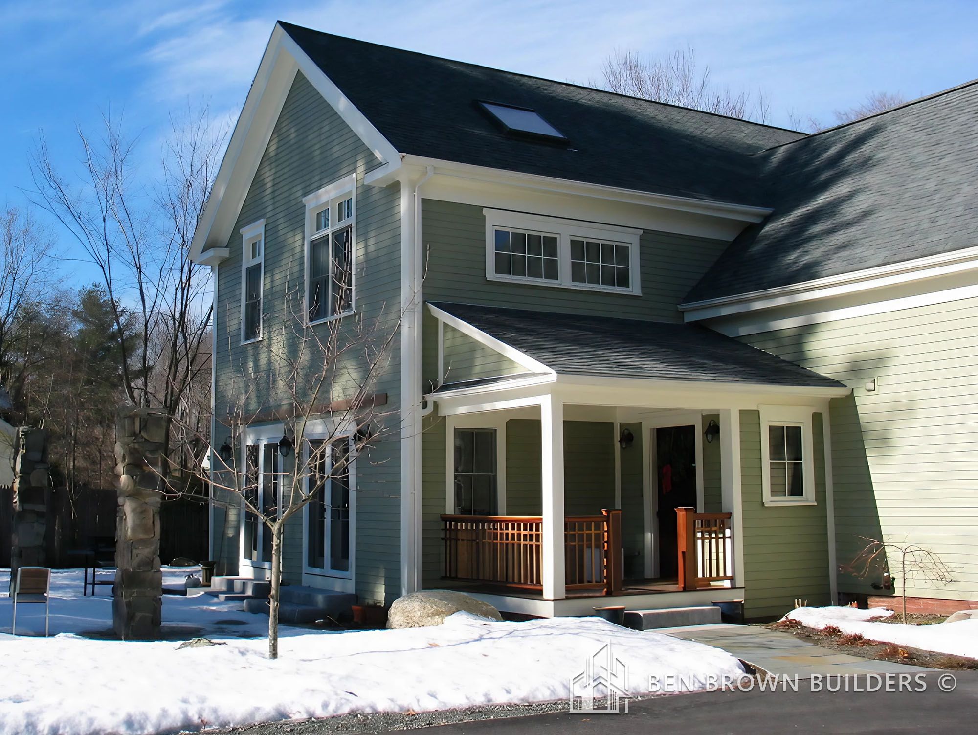 Two-story green house with white trim, a covered porch, and snow on the ground reflecting a clear winter day.