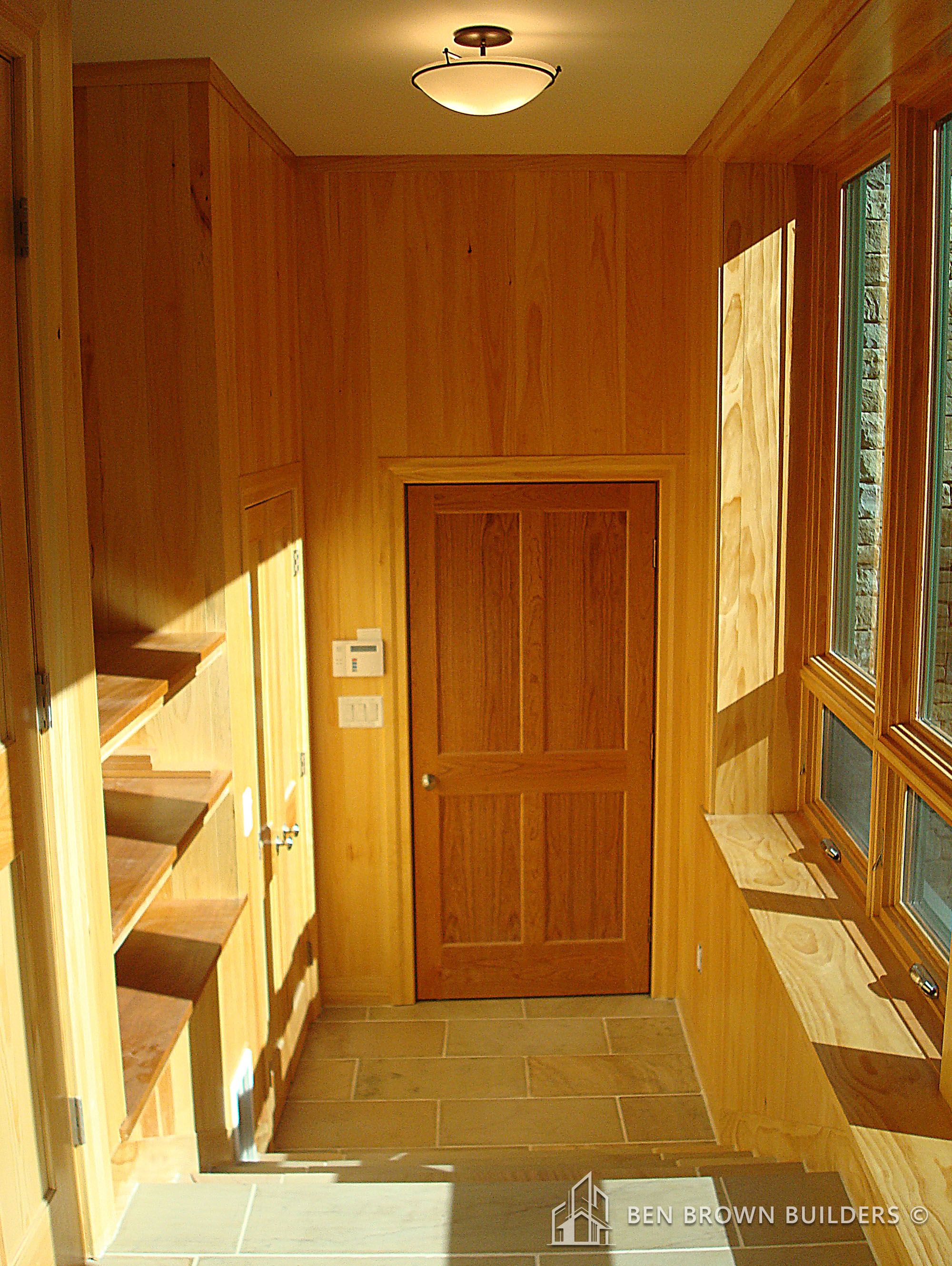 Warmly lit entryway with natural wood paneling, a solid wood door, and tile flooring in a modern home.
