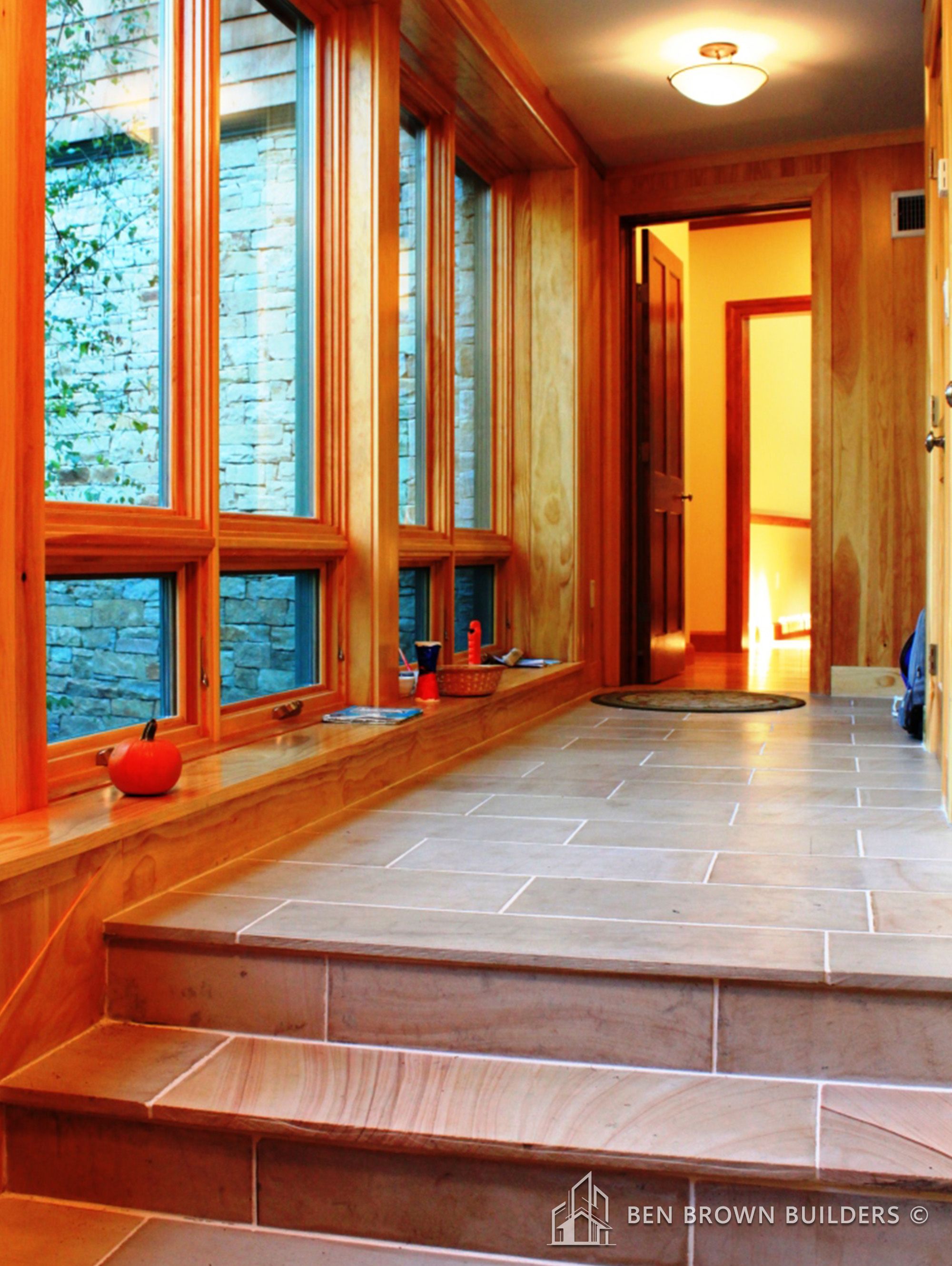 Warmly lit hallway with stone-tiled floors, wooden steps, and large windows showing outdoor greenery