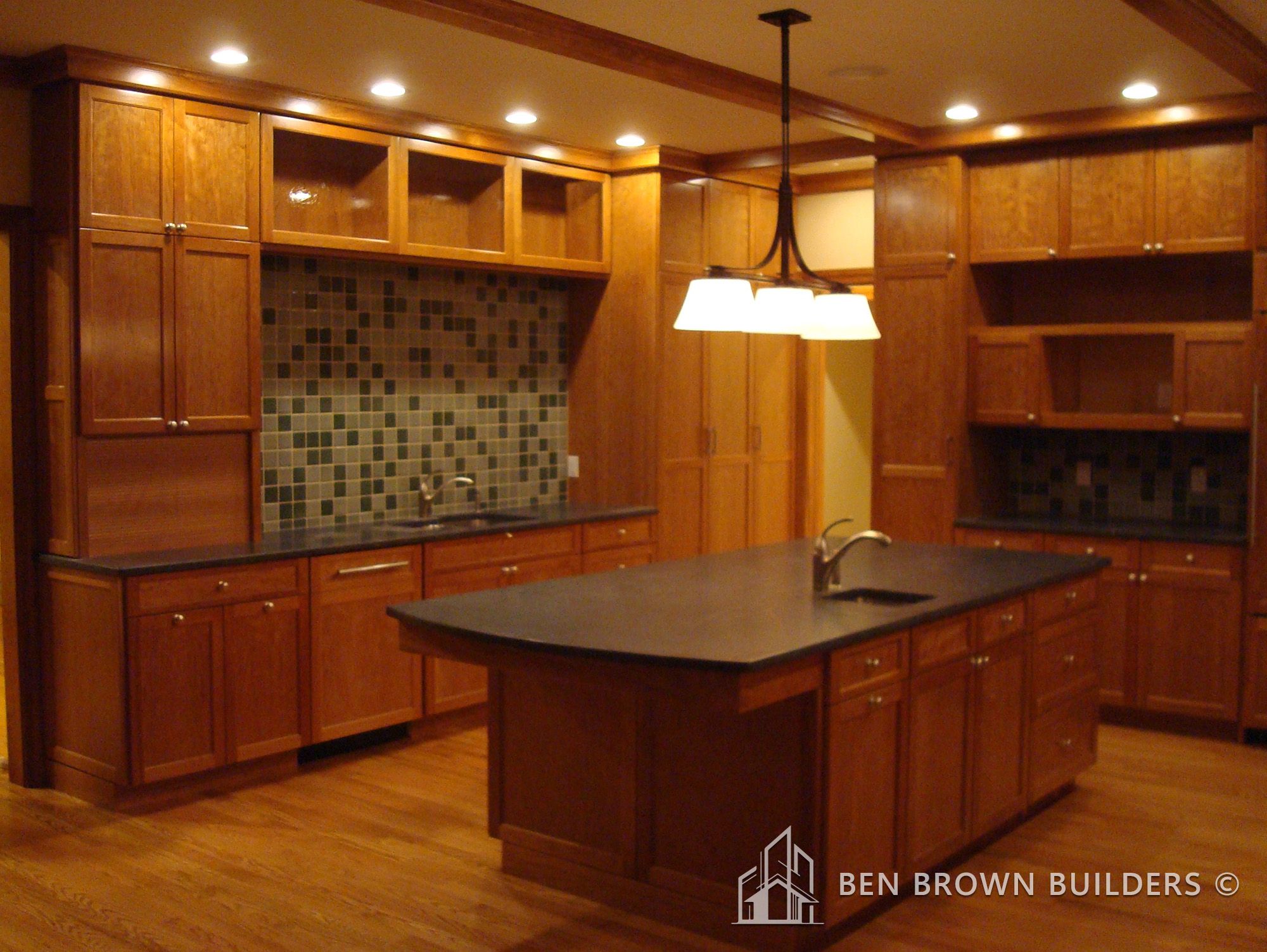 Modern kitchen with cherry wood cabinets, black countertops, a tile backsplash, and warm pendant lighting.