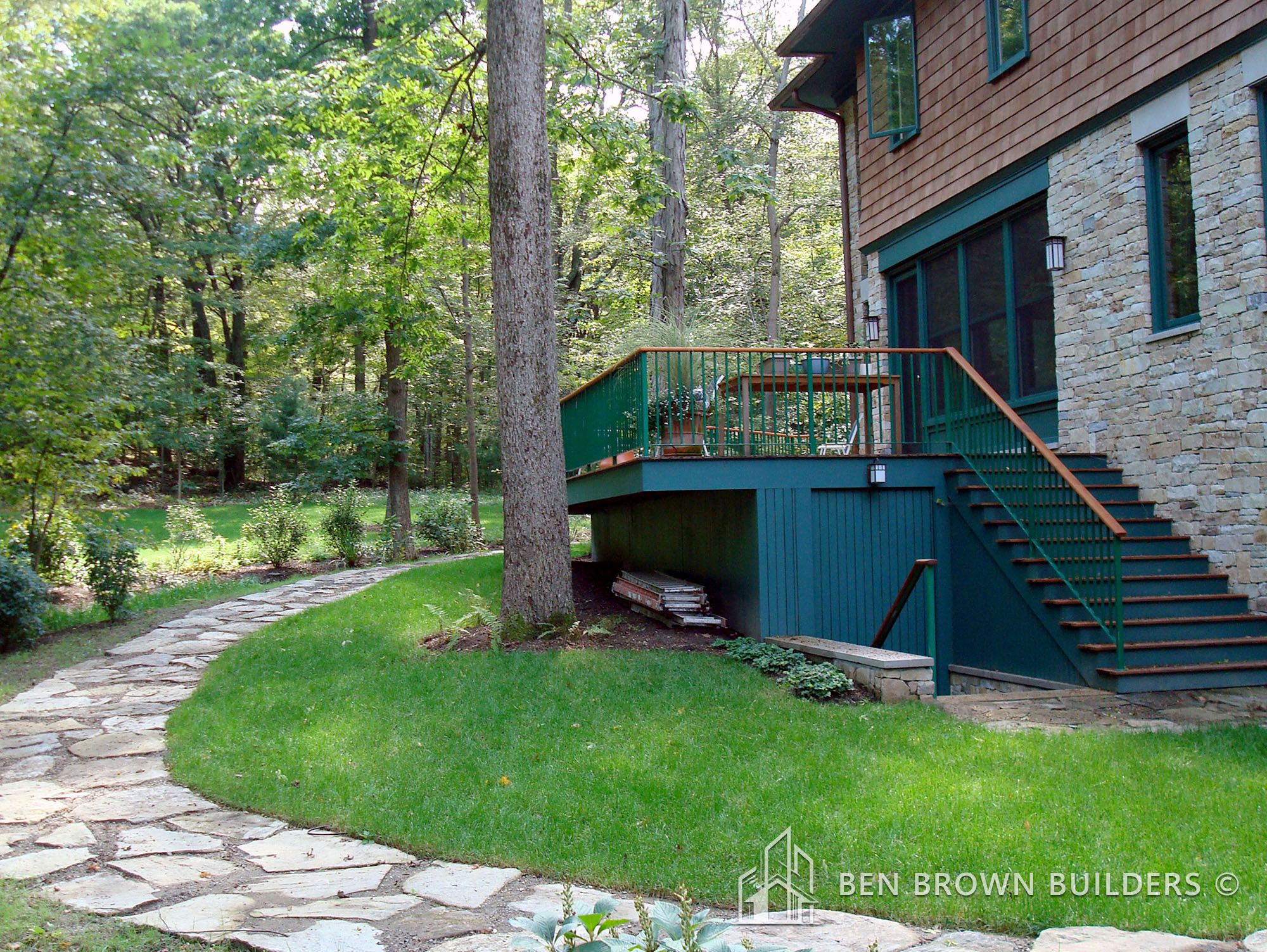 Backyard of a house with stone and siding facade, featuring a raised deck with green railing, and a stone pathway.
