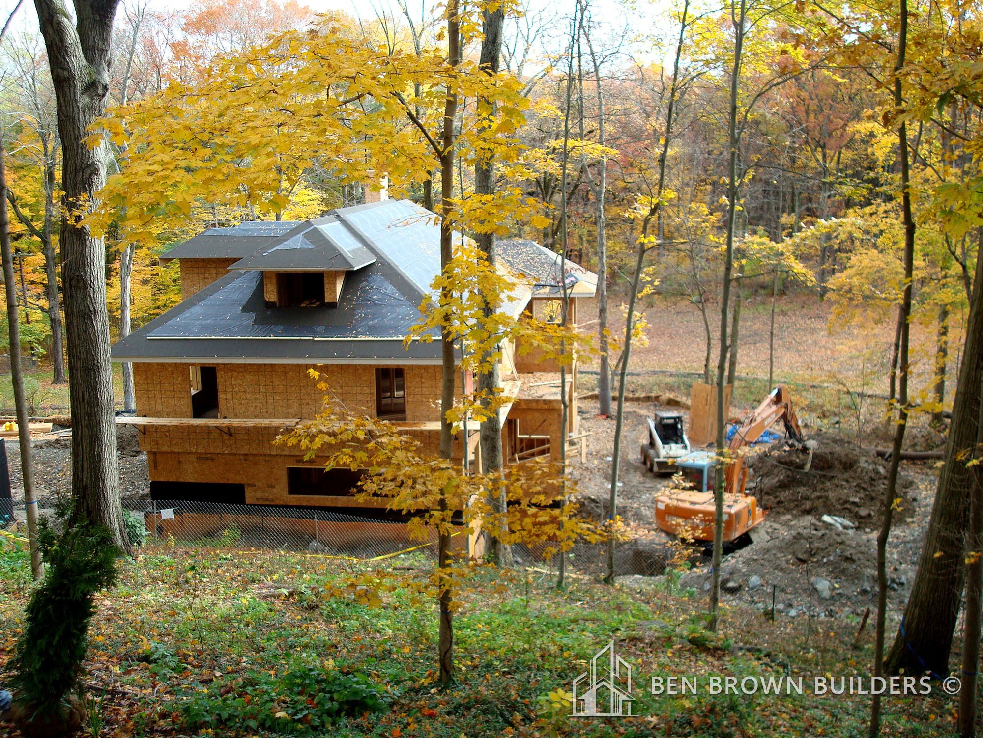 House under construction in autumn with exposed wooden framing, surrounded by trees with yellowing leaves.