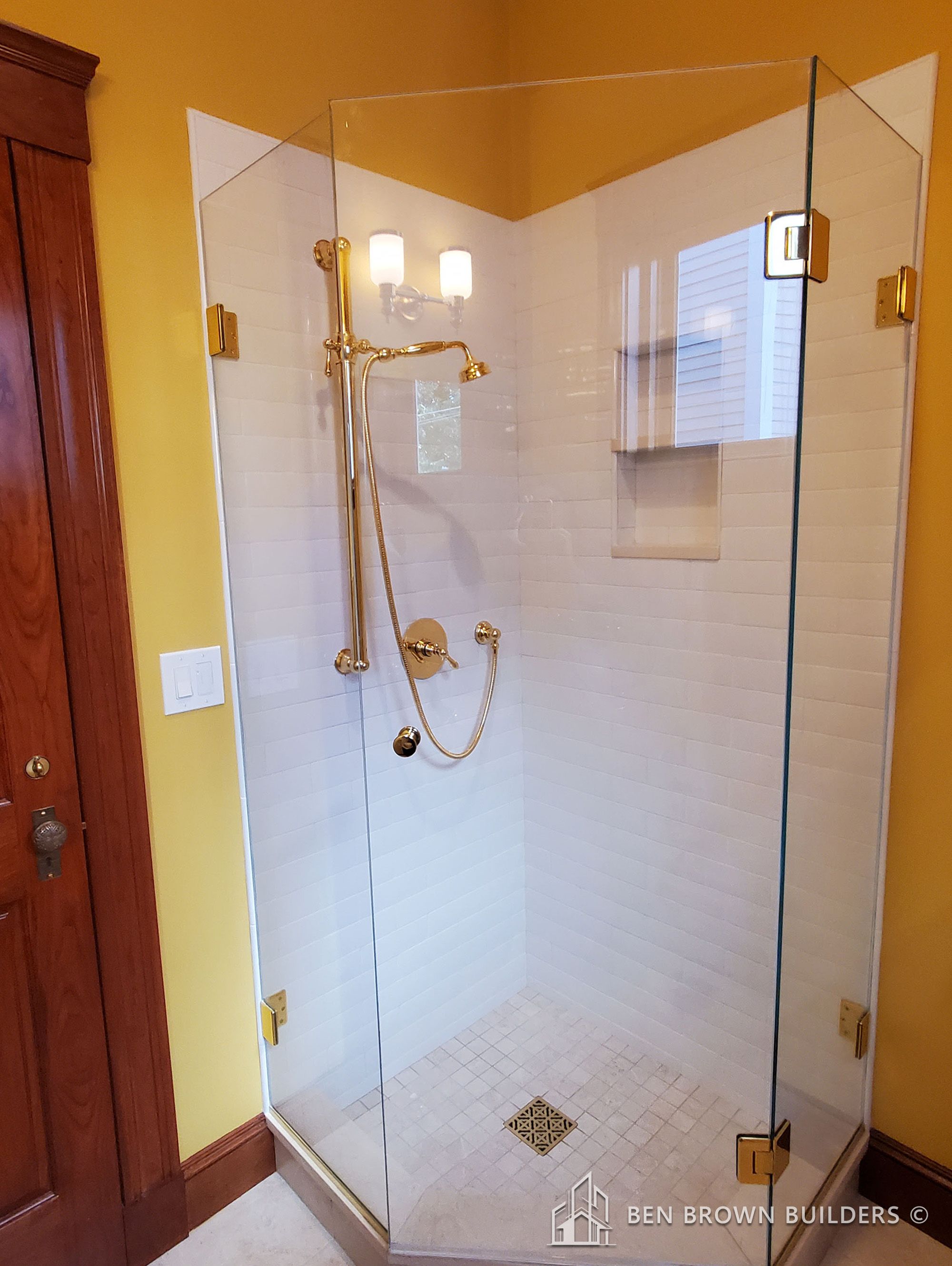 Corner glass shower with white subway tiles, gold fixtures, and a built-in tiled soap dish