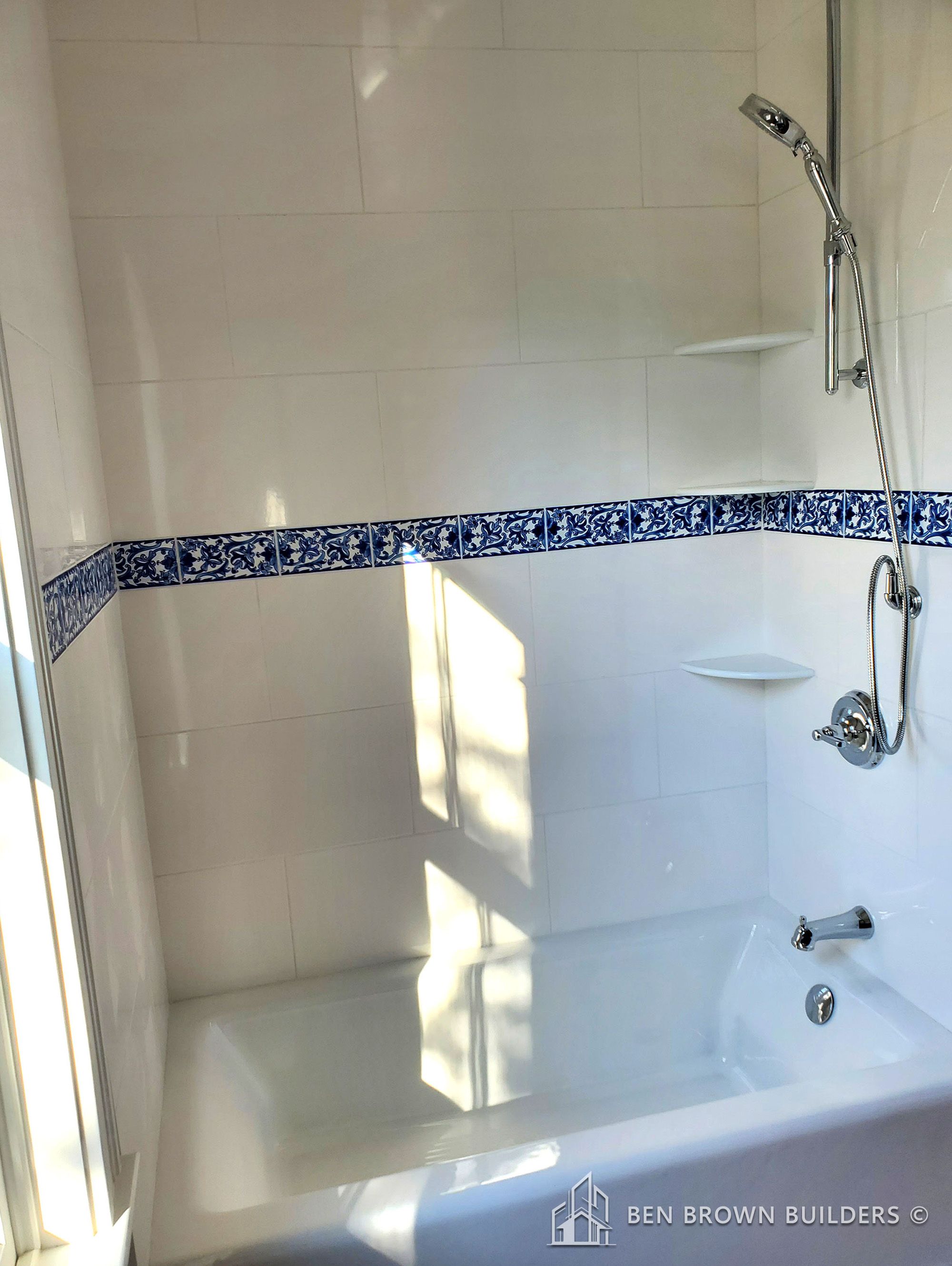 Sunlit bathroom with a white bathtub, blue ornamental tile border, and a handheld shower head