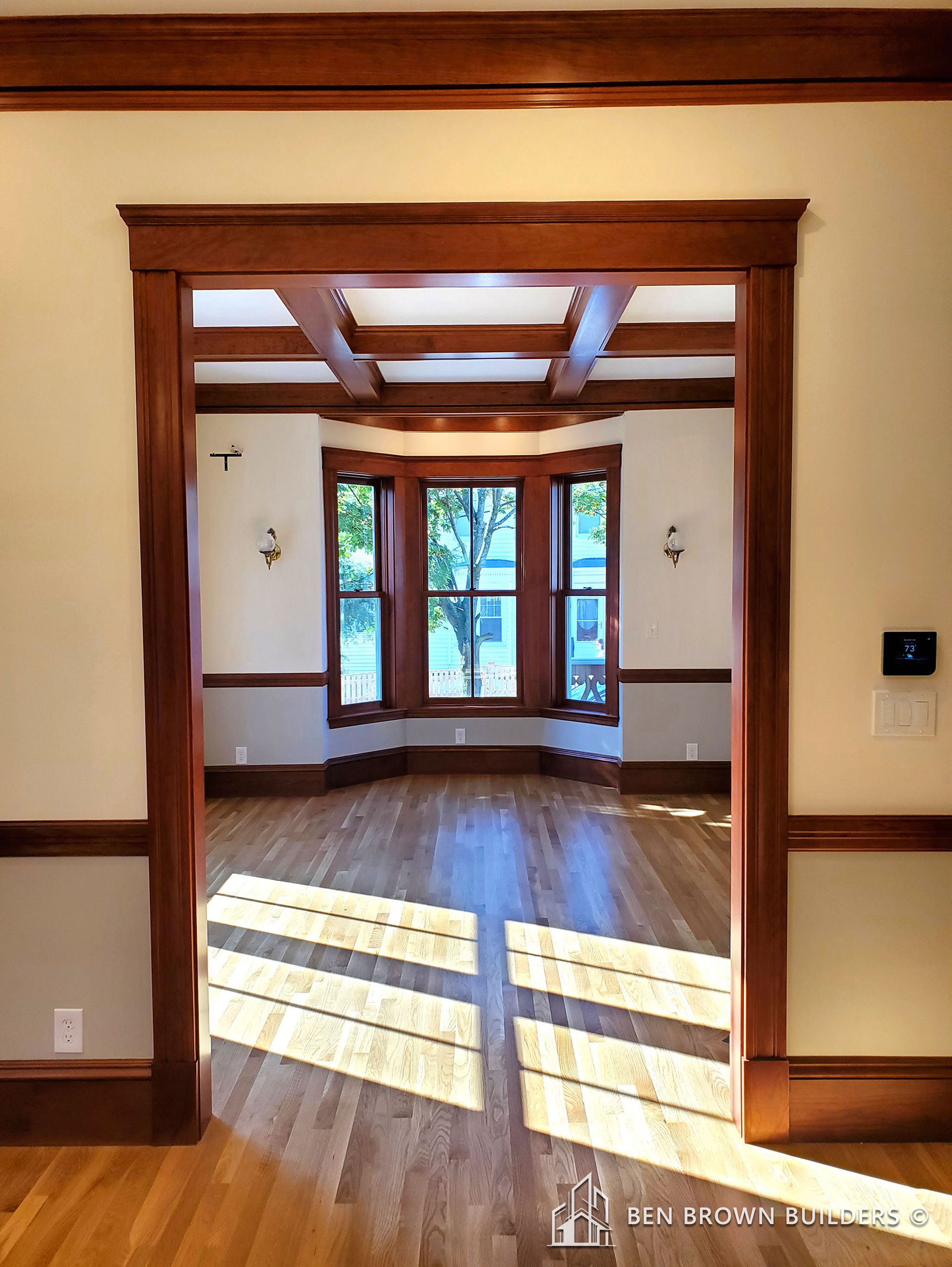 Sunlit room with a bay window, wooden floors, coffered ceiling, and decorative wall sconces