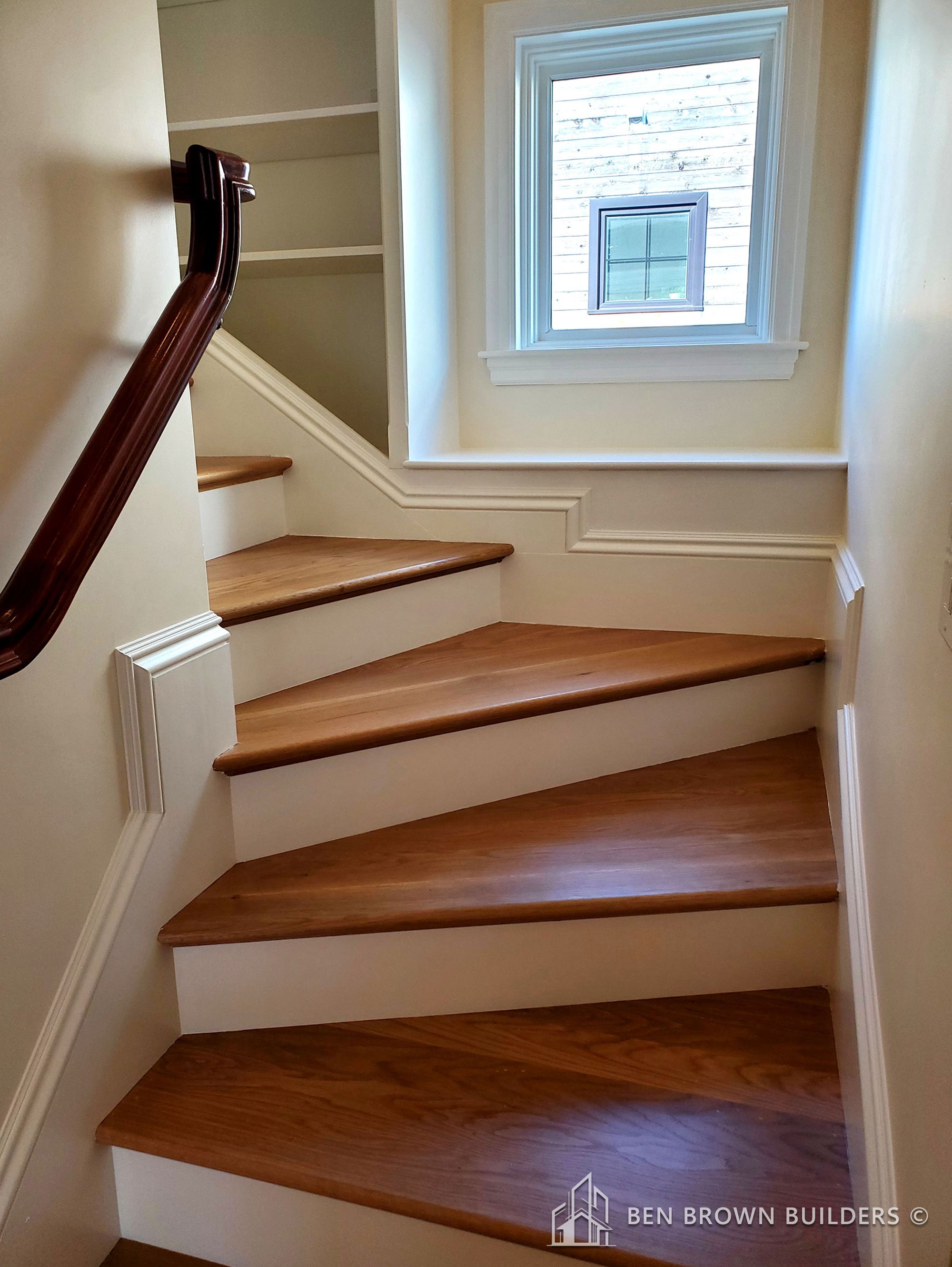 Close-up of a wooden staircase with a rich dark handrail, contrasting with light walls and a small window