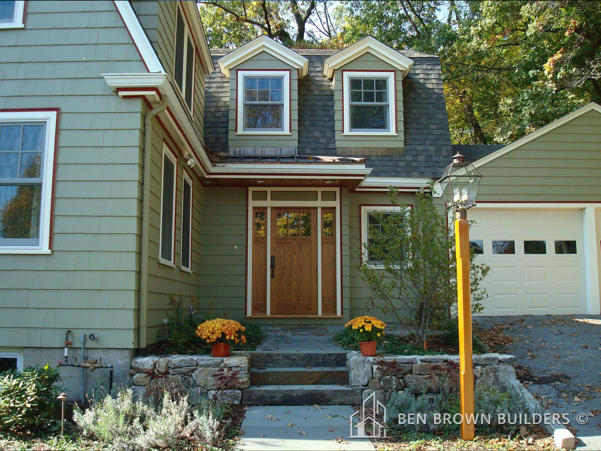 Charming green house with white trim, dormer windows, and an attached garage, featuring autumn decorations