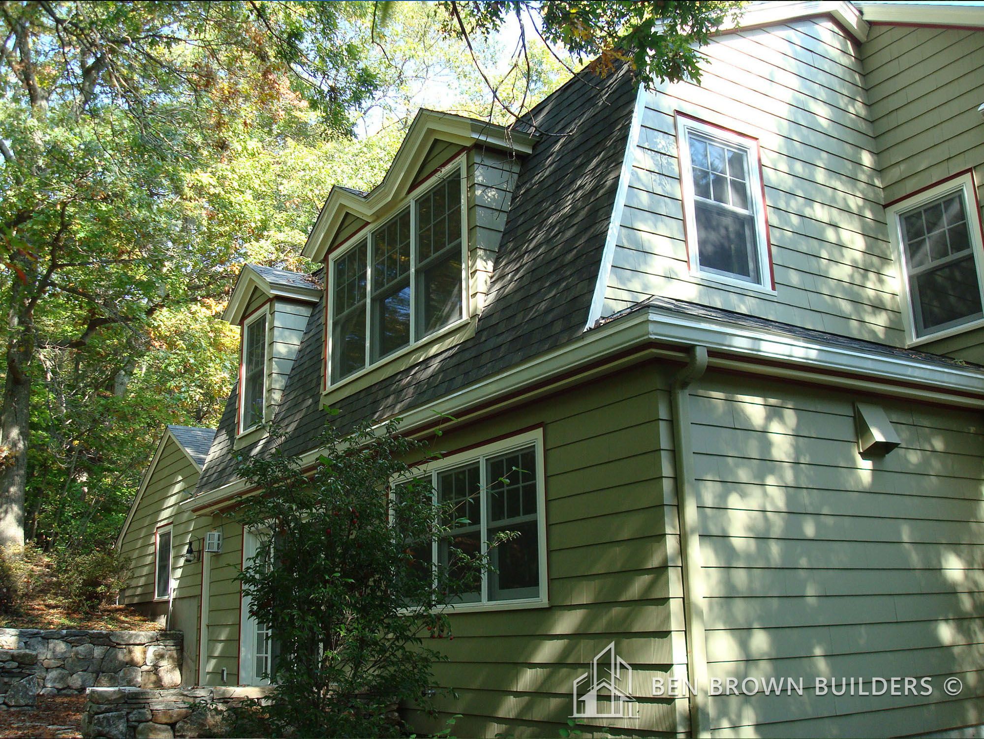 Two-story olive green home with stone steps and surrounding trees casting shadows on the siding